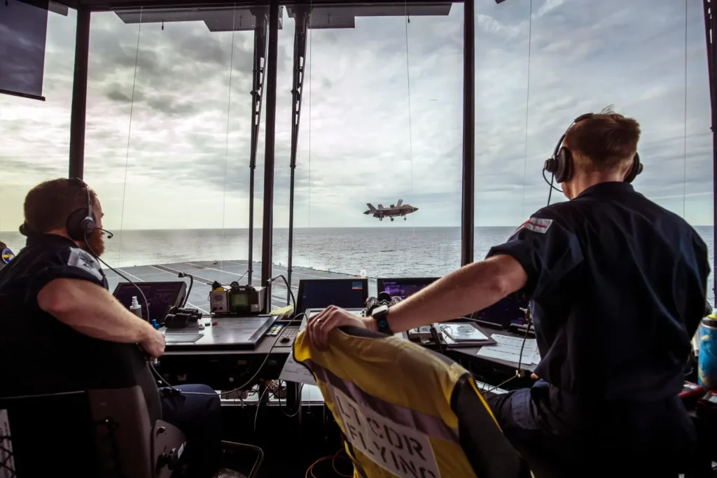 Two operators sat within an air traffic control tower on an air craft carrier with a fighter jet taking off in the background.