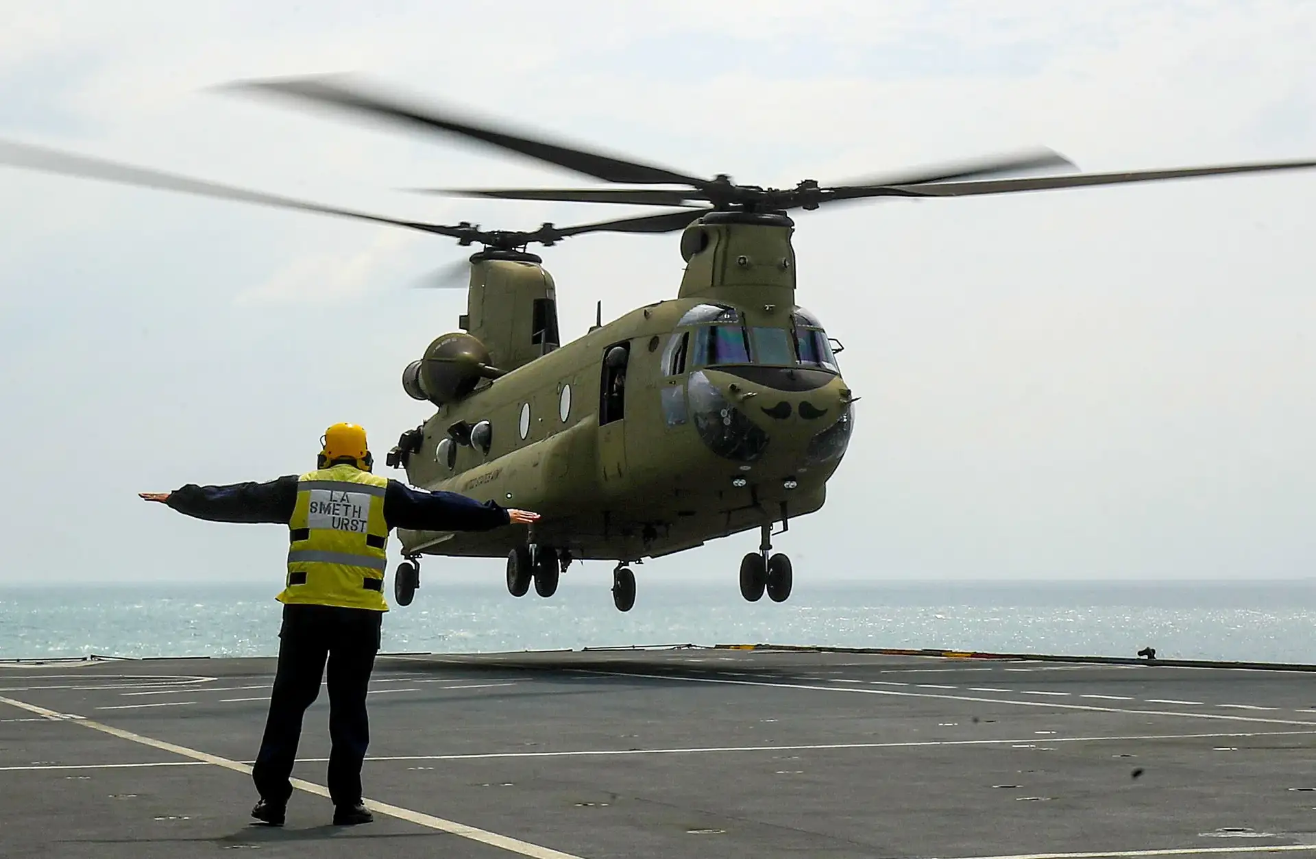 A chinook landing on a military ships deck with an operator signalling to it.