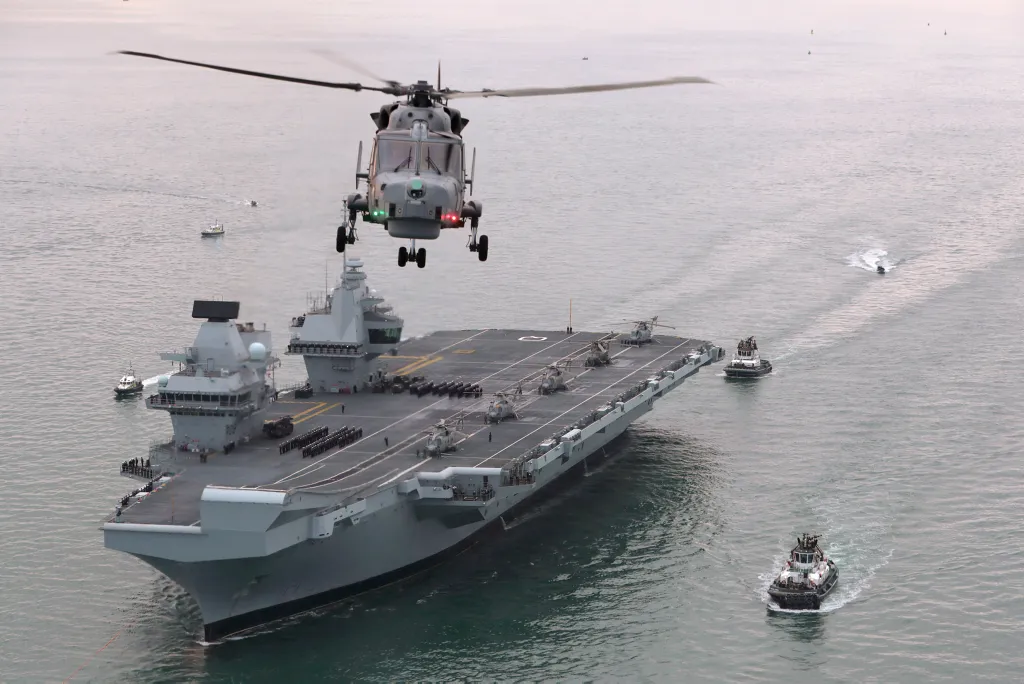 A helicopter having just taken off at sea with an air craft carrier in the background.