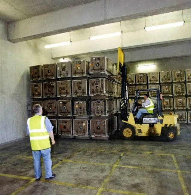 Two workers in a storage facility, one operating a forklift to move products and the other watching him.