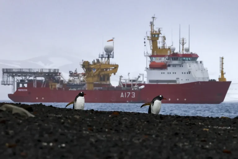 Two penguins walking across a beach with a large cargo ship in the background.