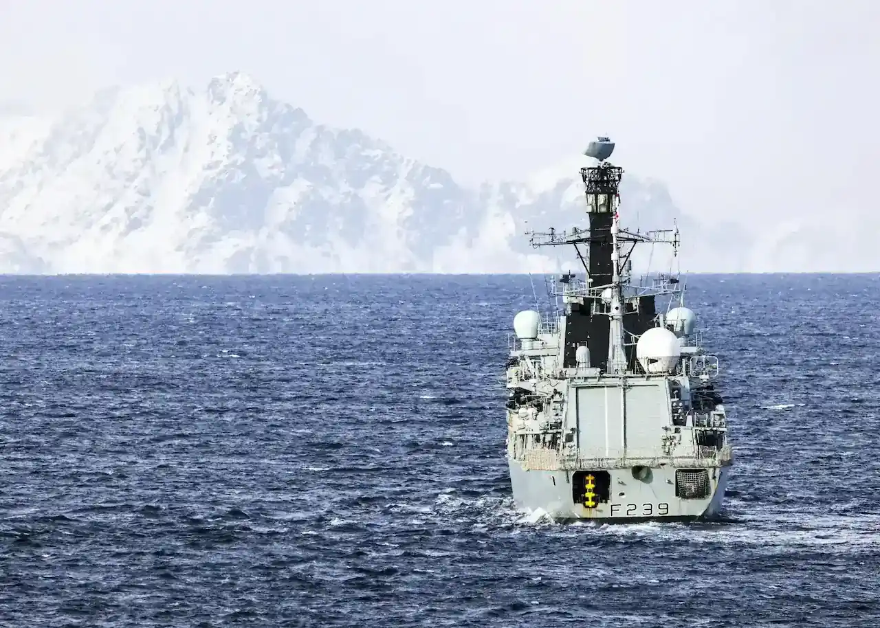 A military ship sailing through the sea with snowy mountains in the background.