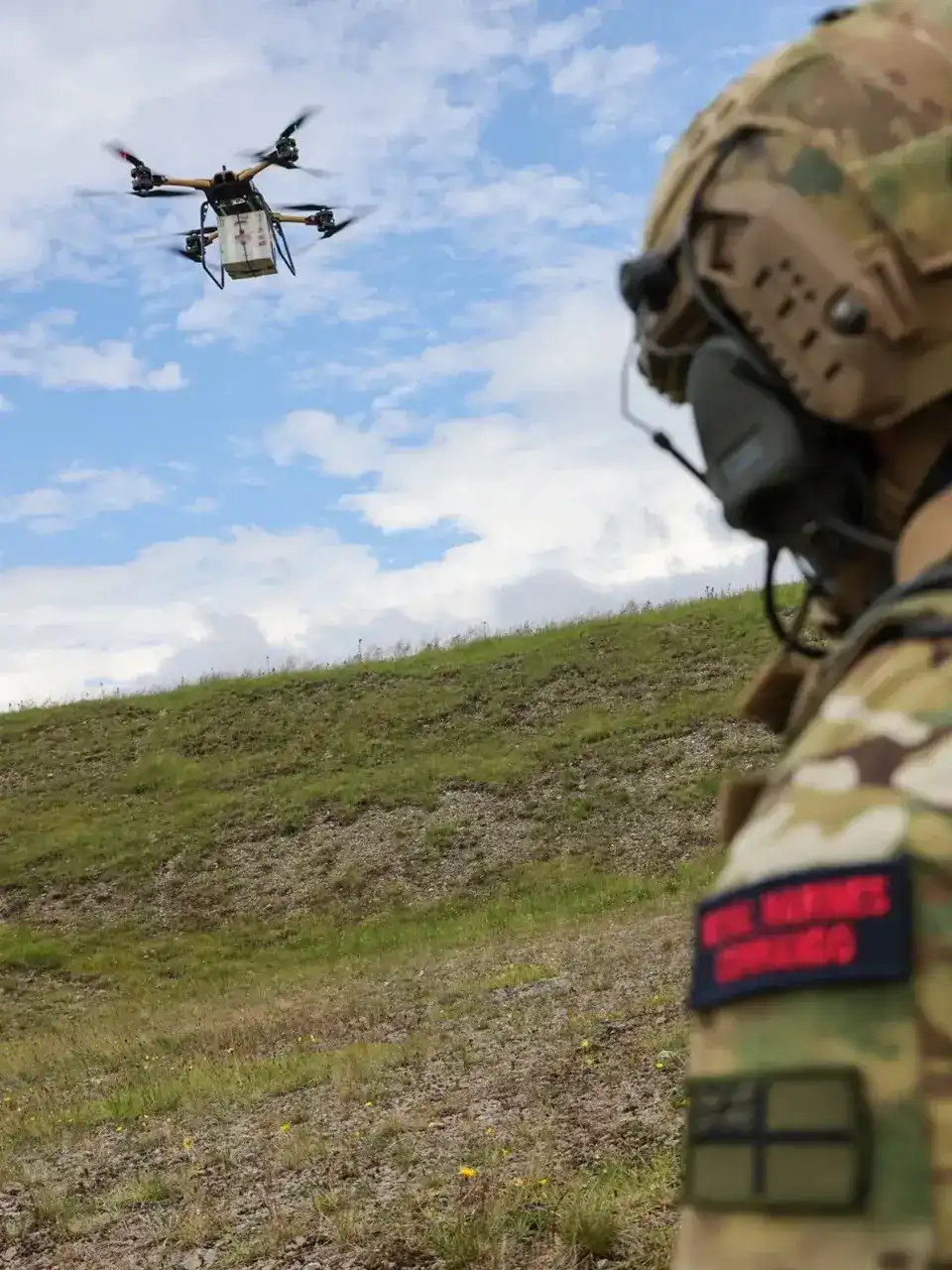 A soldier stood looking at a remotely piloted air system in the air and carrying a package.