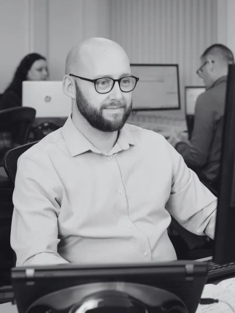 A man in a shirt sat working at a computer in a busy office.