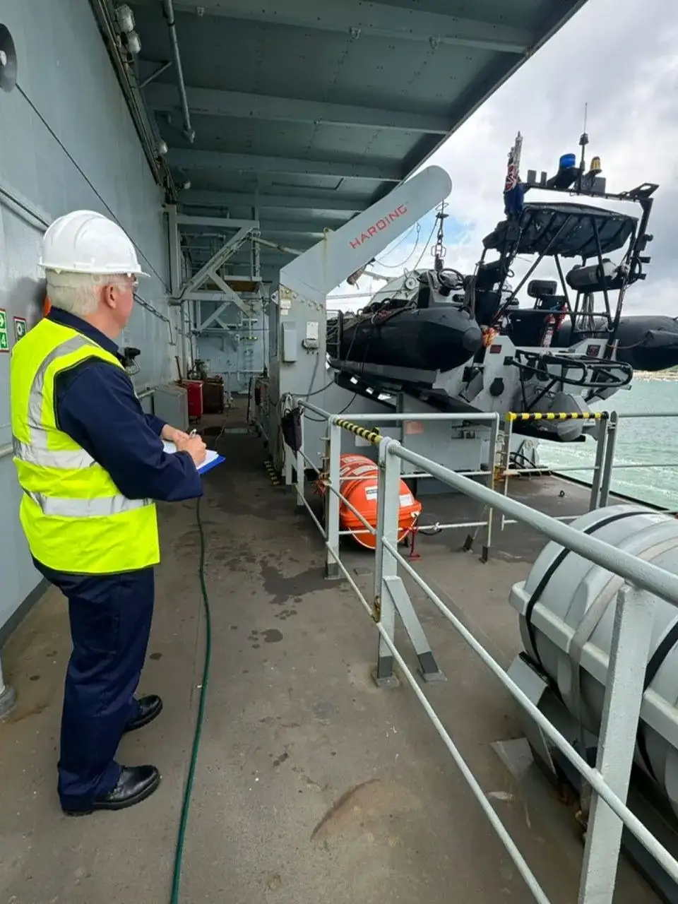 A worker on a ship, writing on a clipboard and observing a deployable RIB.