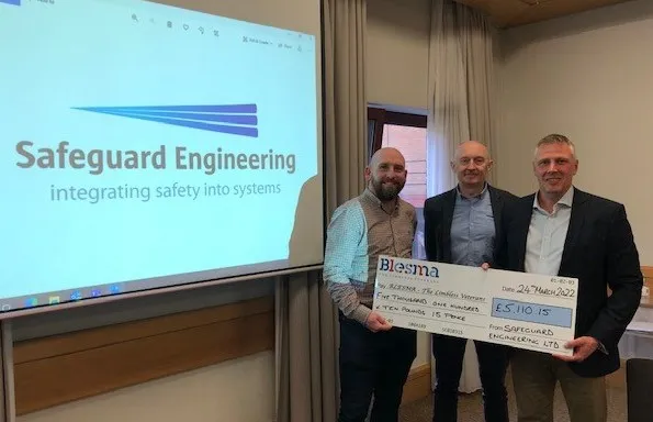 Three people stood side-by-side holding a cheque next to a projector screen with the Safeguard Engineering logo on it.