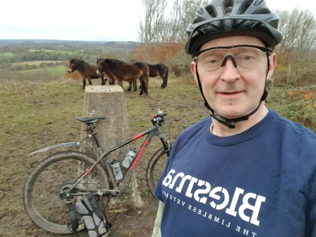 A man wearing a cycling helmet, glasses, and Blesma t-shirt with a bike and wild horses in the background.