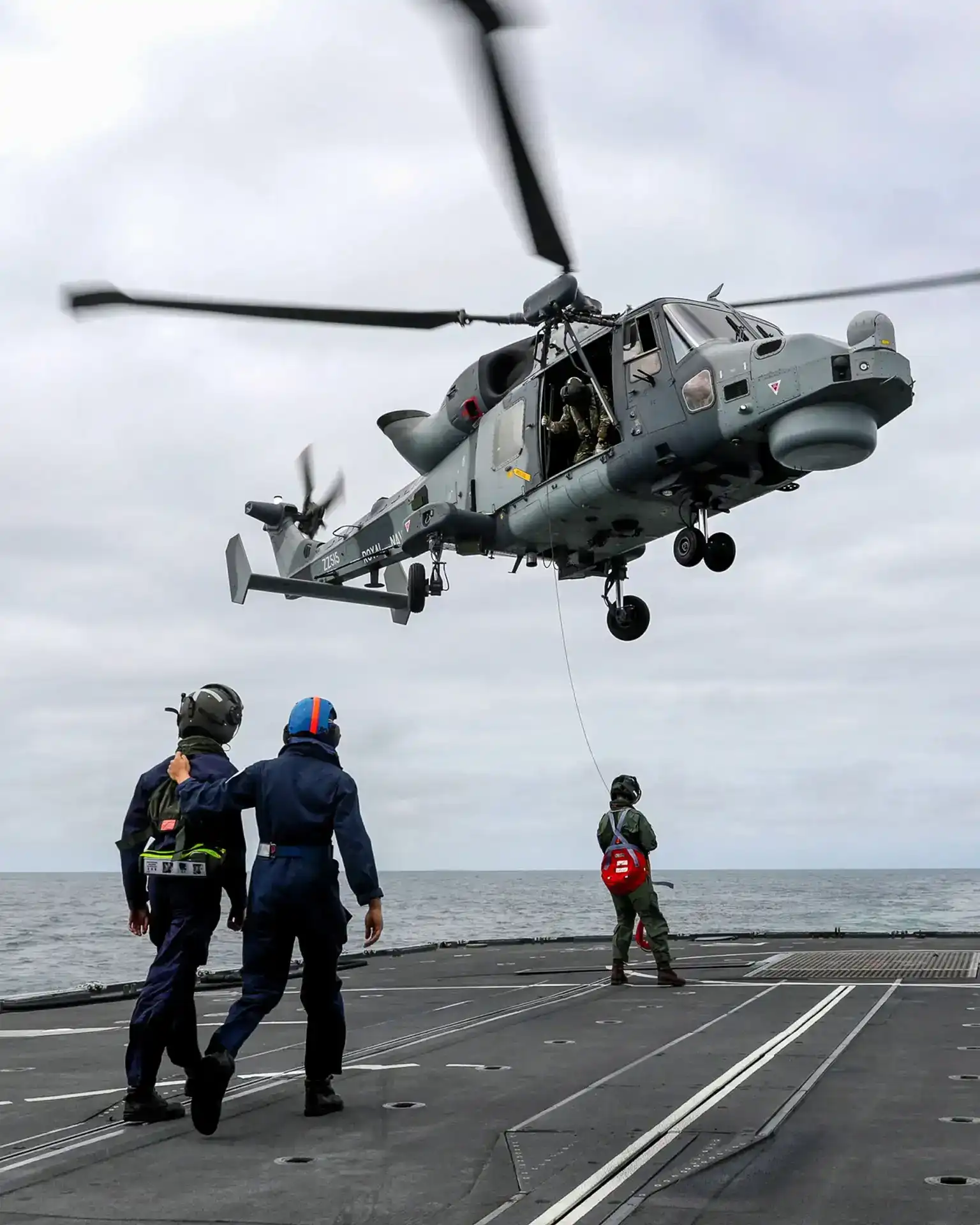 A helicopter landing on a flight deck with three operators preparing for its landing.