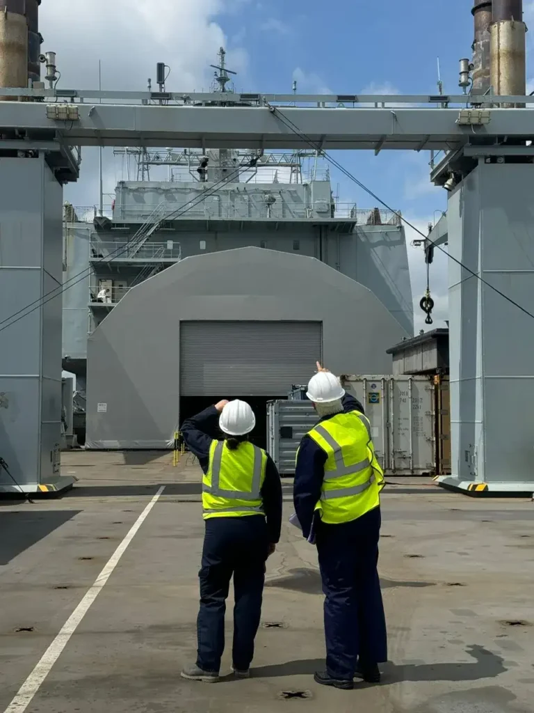 Two workers stood on a ships deck pointing and looking at machinery.