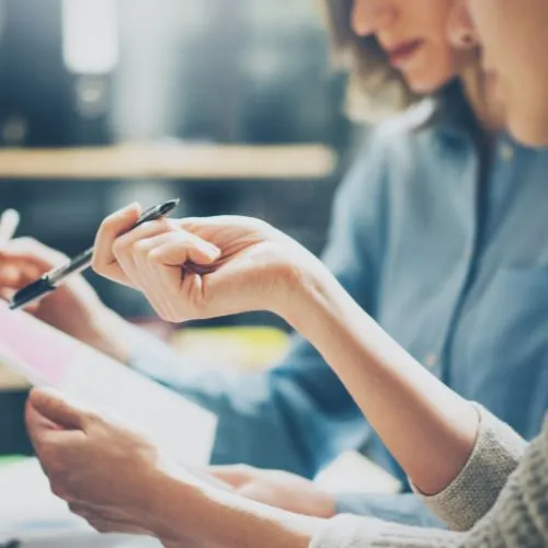 Two people sat at a desk, looking at paper work and pointing to words with a pen.